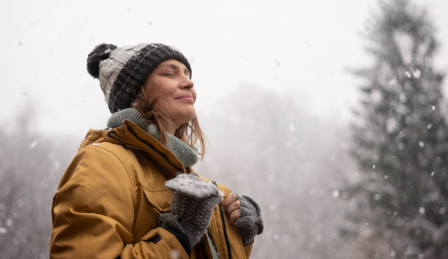 Woman outside smiling at falling snow with evergreens in the background